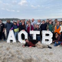 Agile on the Beach conference attendees at the beach party, gathering around the big letters "AOTB" in front of the water